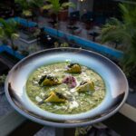 A dish of spinach curry with paneer dumplings garnished with flowers, served outdoors in a tropical setting.