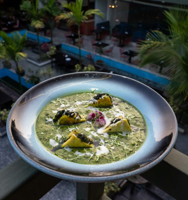 A dish of spinach curry with paneer dumplings garnished with flowers, served outdoors in a tropical setting.